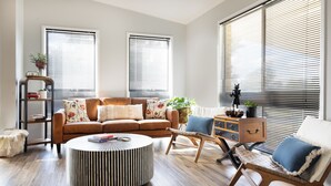 A modern living room with a brown sofa, a round coffee table, and a wooden chair. There are two windows with blinds, a bookshelf, and a small table with a plant.