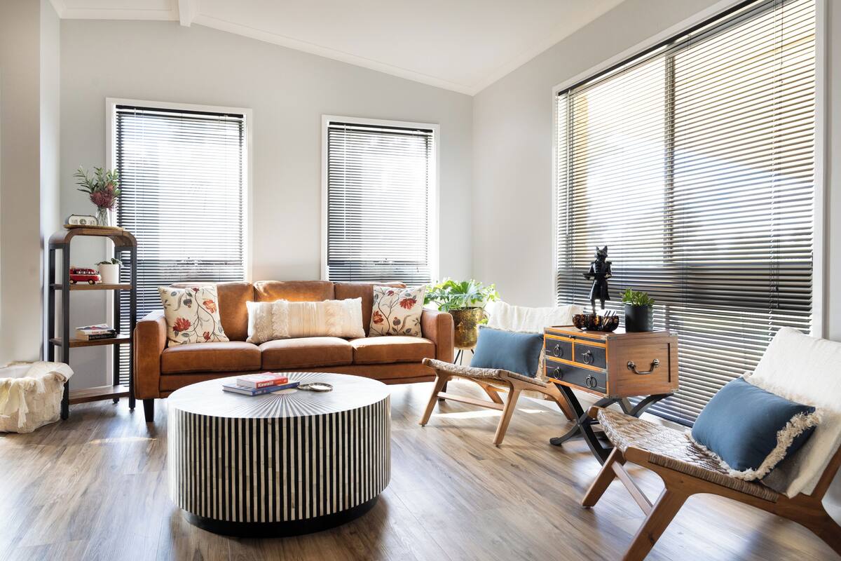 A modern living room with a brown sofa, a round coffee table, and a wooden chair. There are two windows with blinds, a bookshelf, and a small table with a plant.