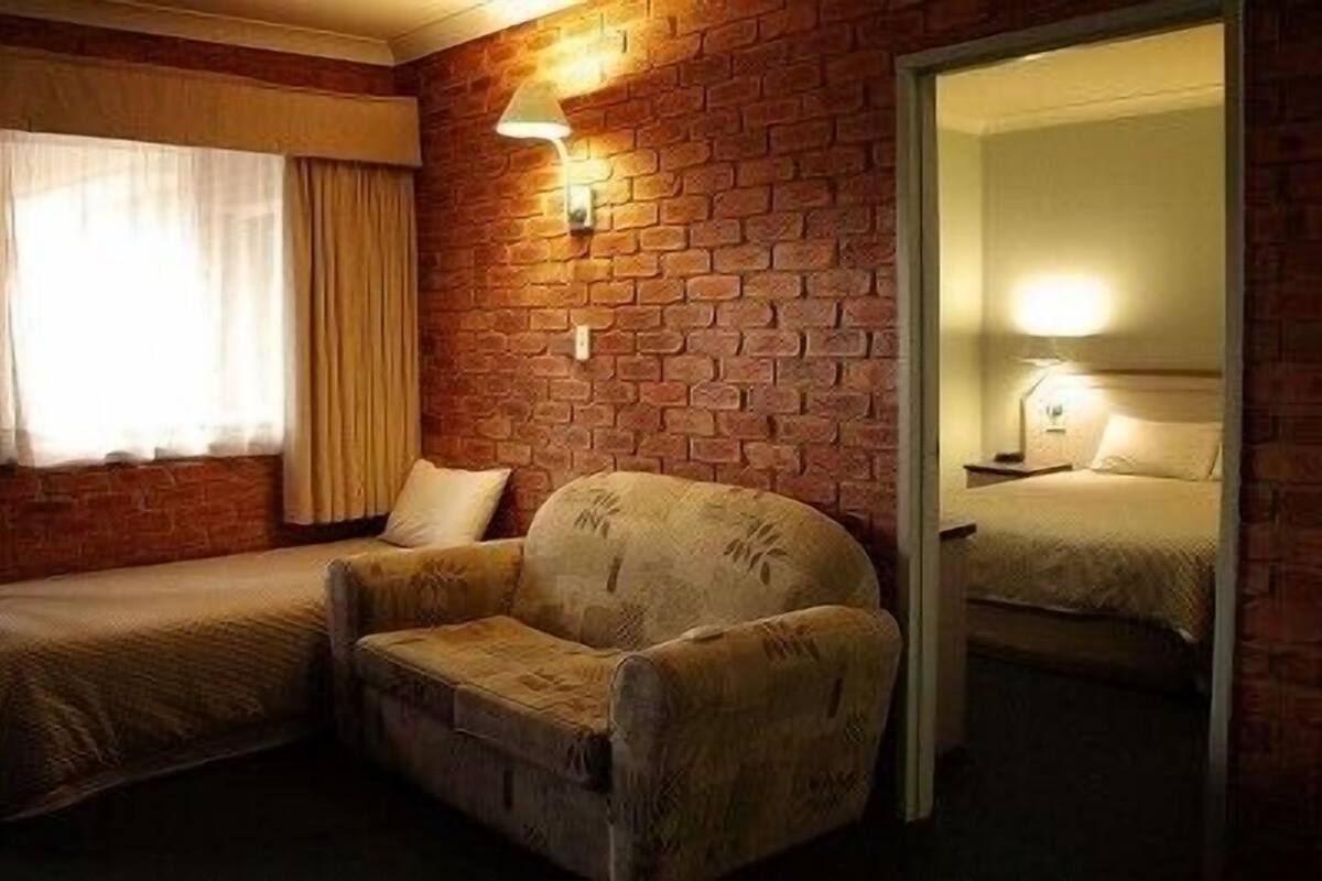 A room with a brick wall, a single bed, a chair, and a window with curtains.