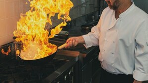 A chef in a white shirt cooking with a pan on a stove with flames.