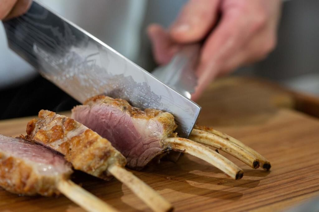 A person cutting a cooked lamb rack on a wooden cutting board.