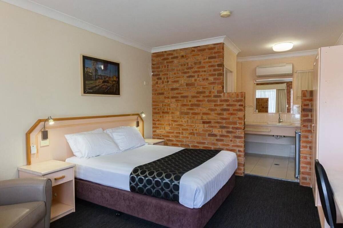 A hotel room with a brick accent wall, a bed with white linens, a nightstand with a lamp, a desk, and a mirror.