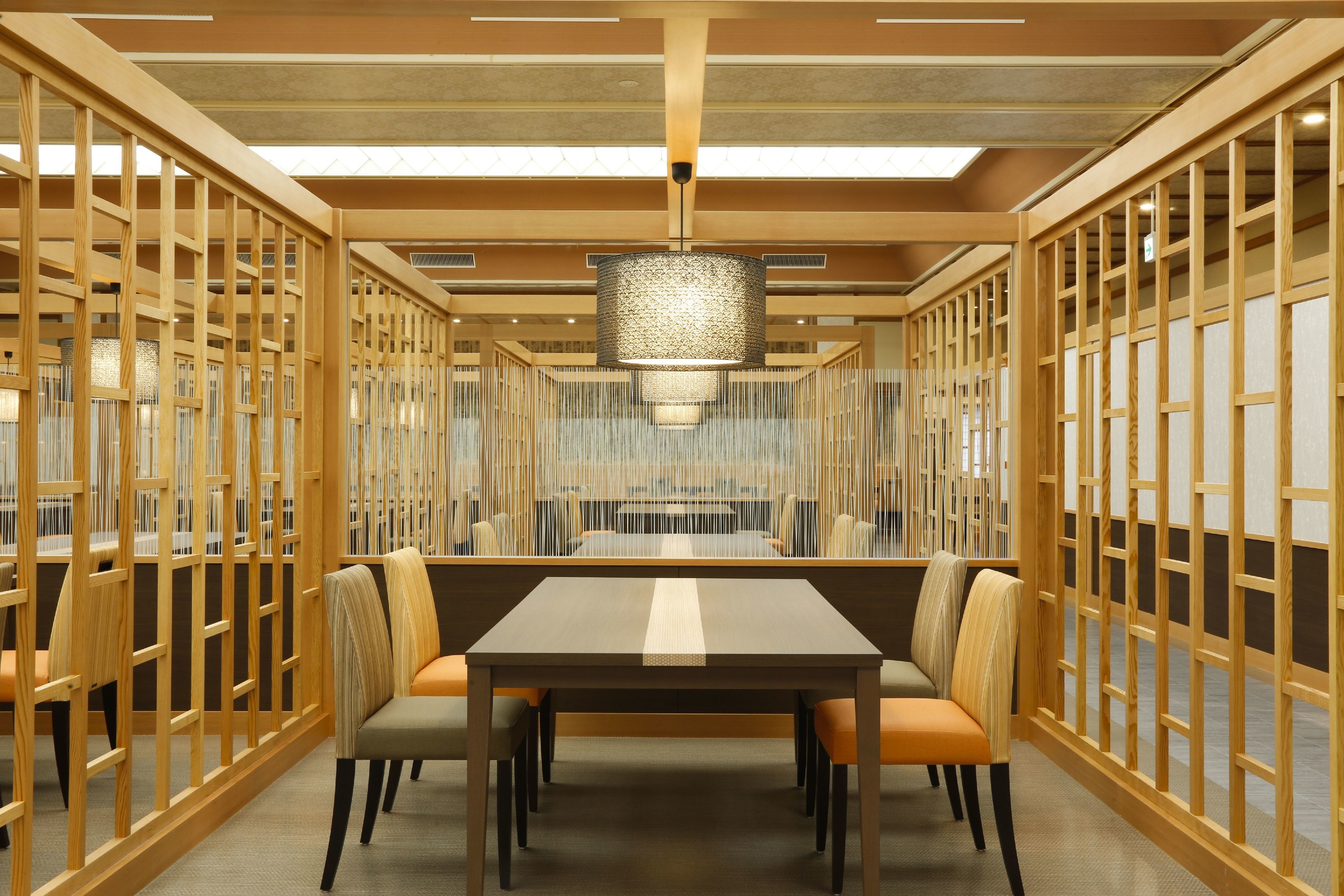 A modern dining area with a long table and chairs, wooden partitions, and a hanging light fixture.