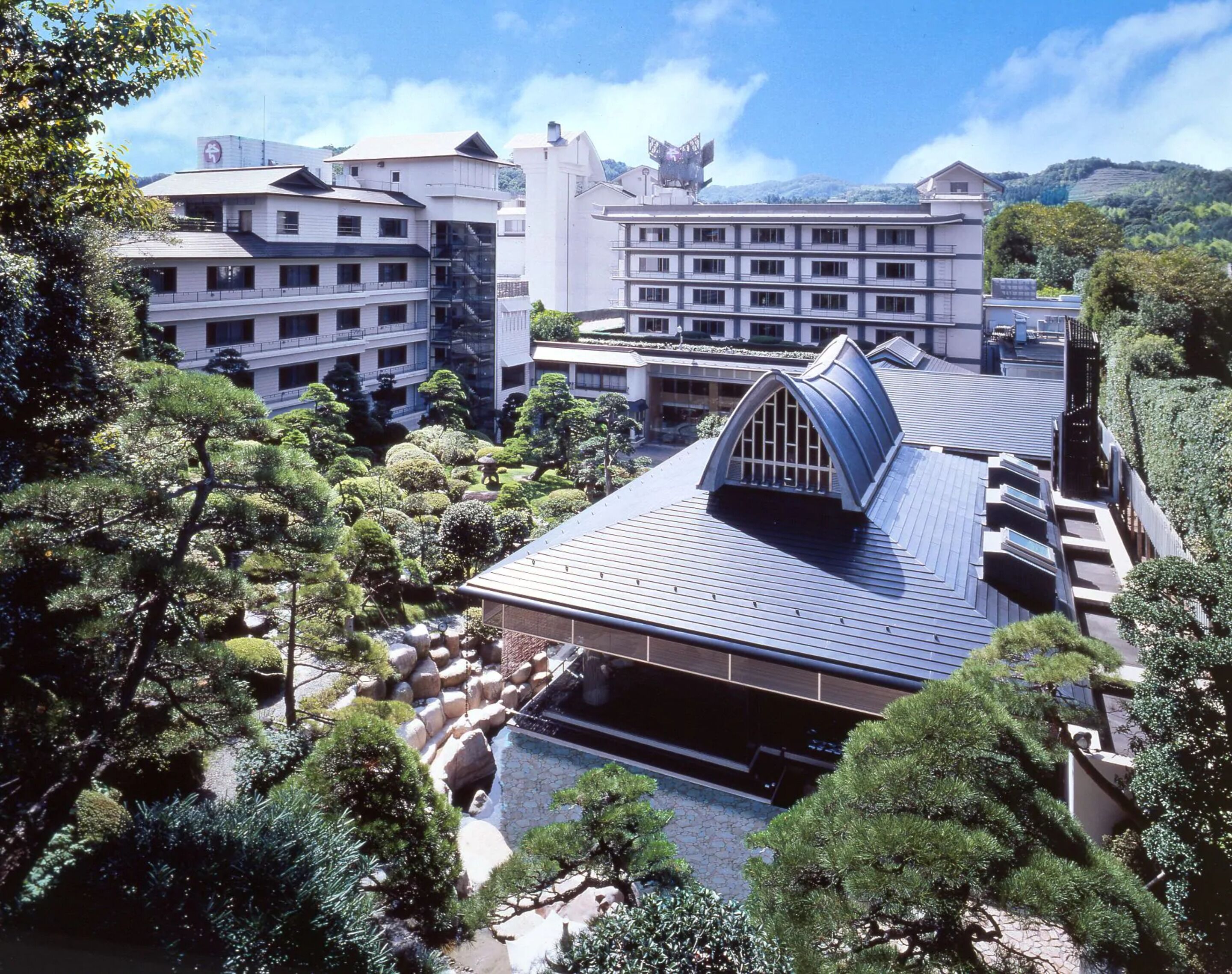 A hotel with a traditional Japanese architectural style, featuring a prominent curved roof and a garden with neatly trimmed trees and a stone pathway.