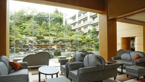 A modern lounge area with sofas and a small table, large windows offering a view of a landscaped garden and a building in the background. - Tamatsukuri Grand Hotel Choseikaku (Matsue)