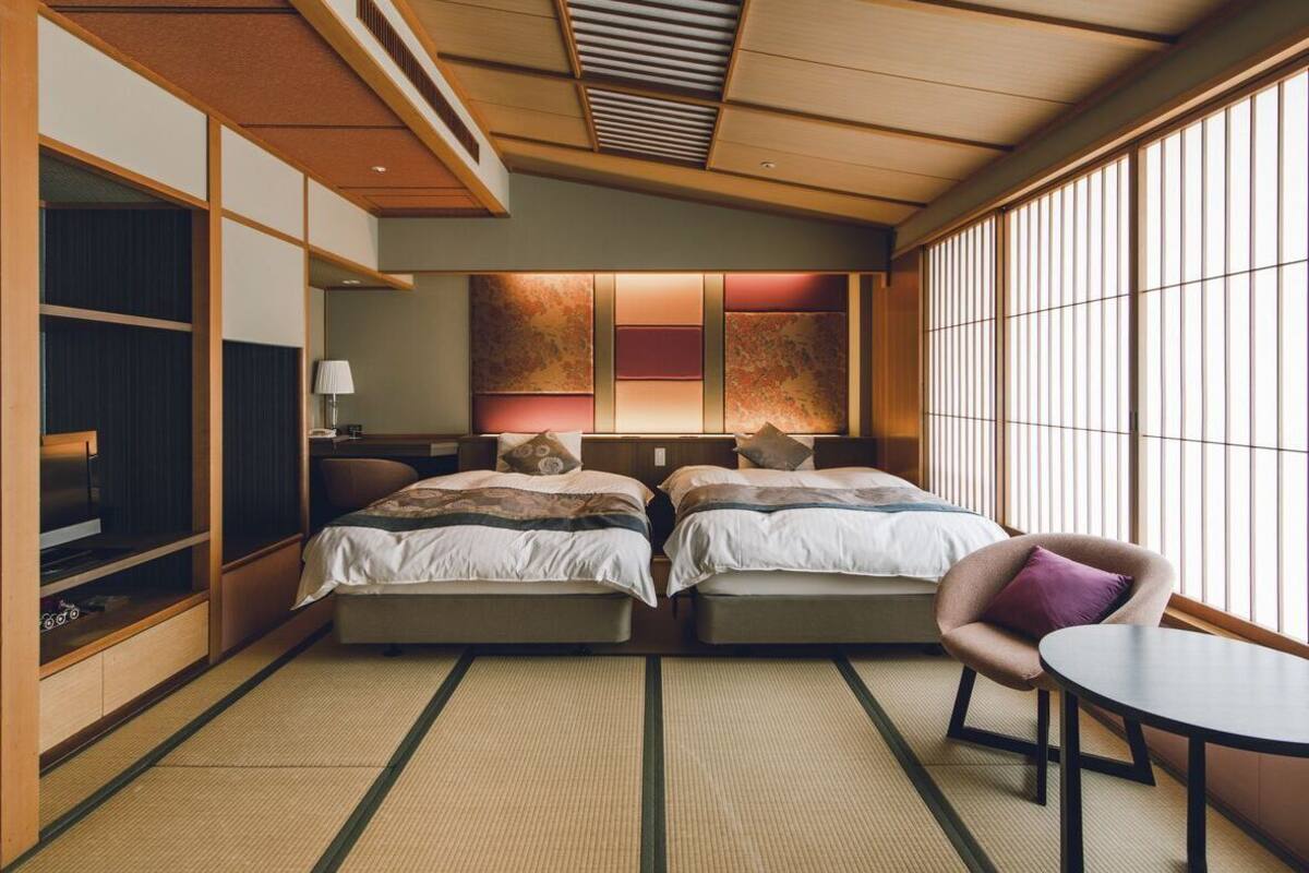 A traditional Japanese-style hotel room with tatami flooring, two beds, a television, a small table, and a window with vertical blinds.