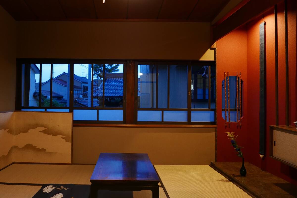 Une pièce de style japonais traditionnel, avec un sol recouvert de tatamis, un paravent en papier de riz (shoji), une petite table basse et une vue sur un quartier résidentiel par la fenêtre.
