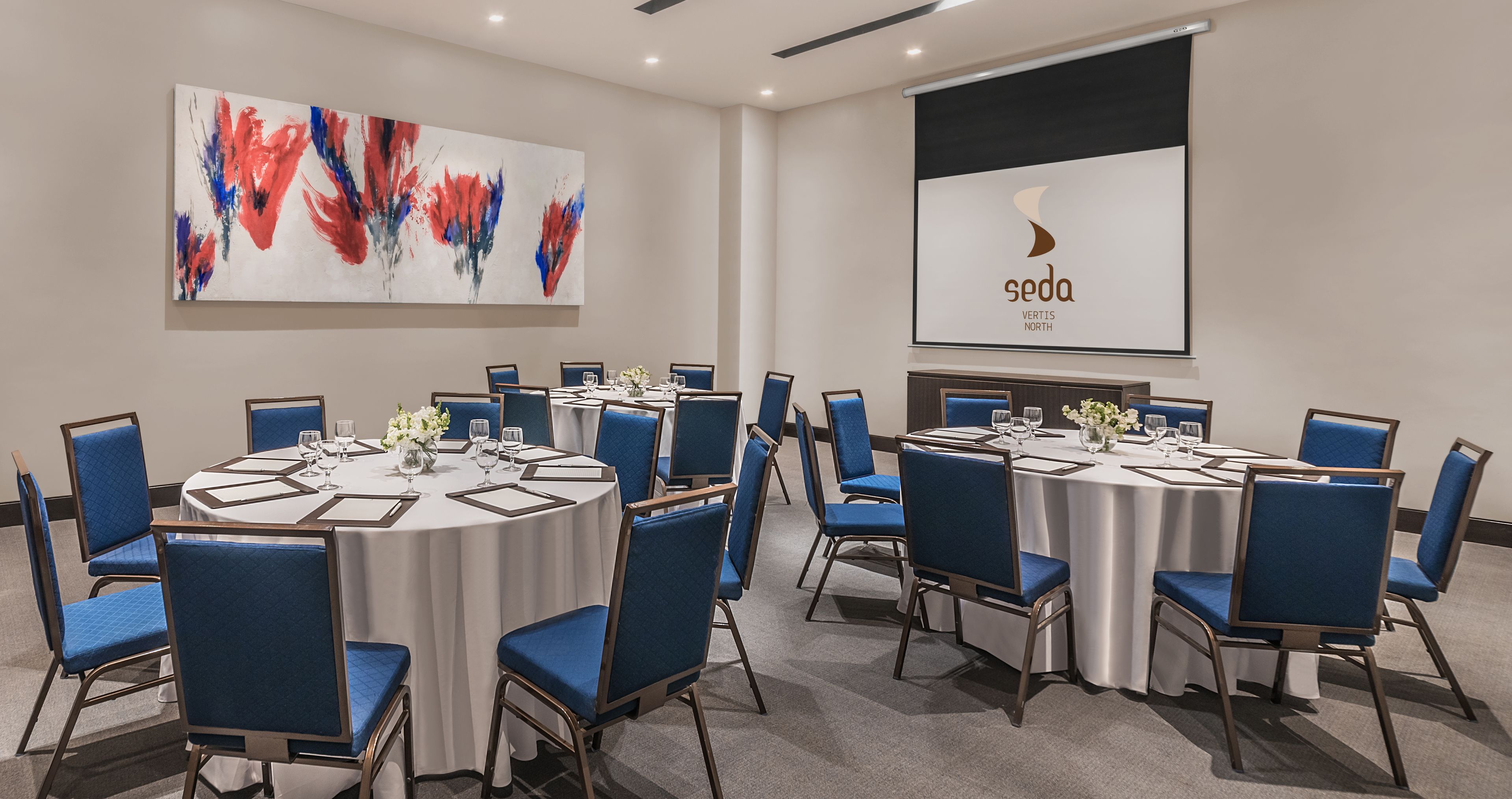 A conference room with round tables, blue chairs, and a projector screen displaying 'seda Vertis North'.