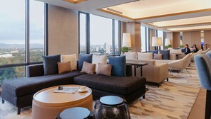 A modern hotel lobby with a large sectional sofa, round coffee table, and a view of the cityscape through large windows.