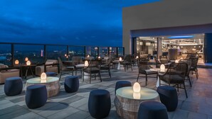 A rooftop bar with outdoor seating, including round tables and chairs, and a city view.