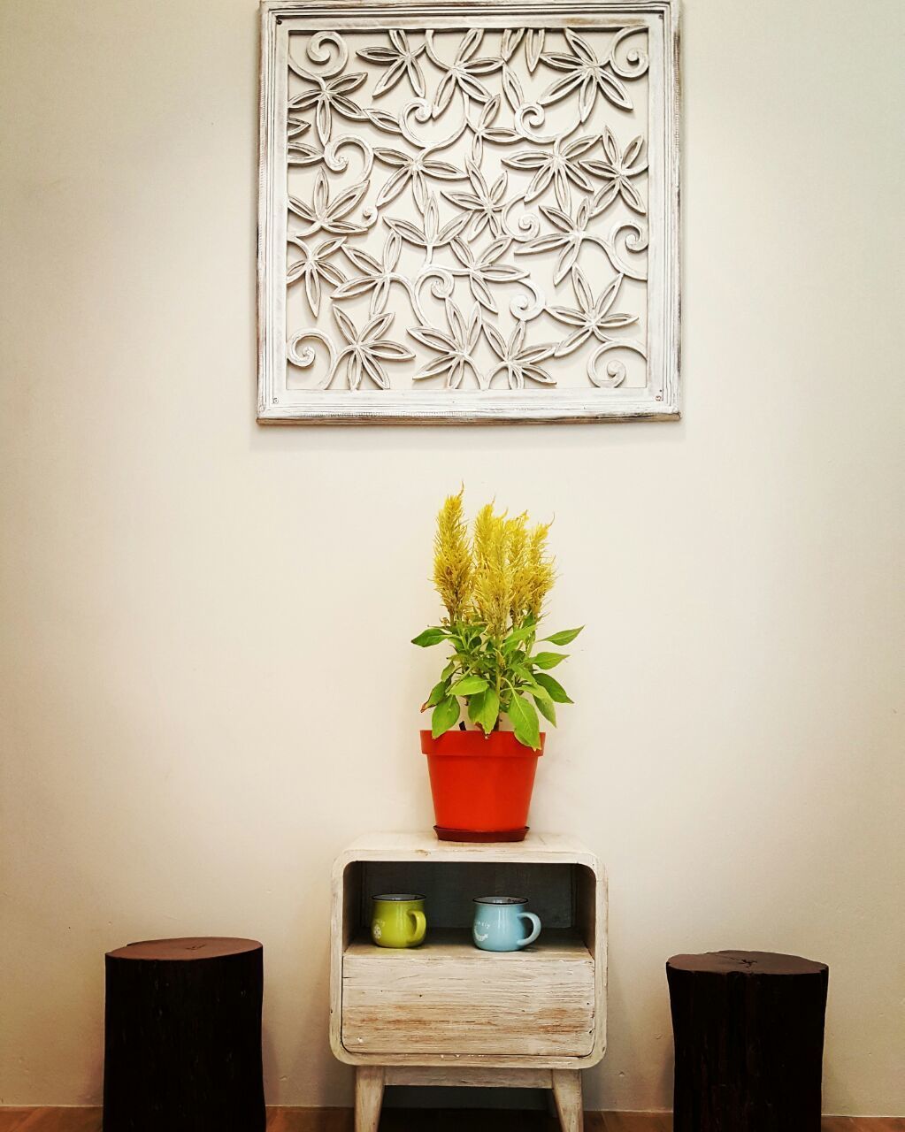 A wall-mounted decorative panel, a potted plant, and a small wooden shelf with two mugs.