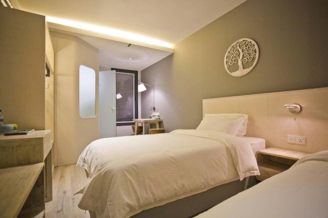 A hotel room with two beds, a desk, and a chair. There is a door leading to another room and a wall-mounted light fixture.