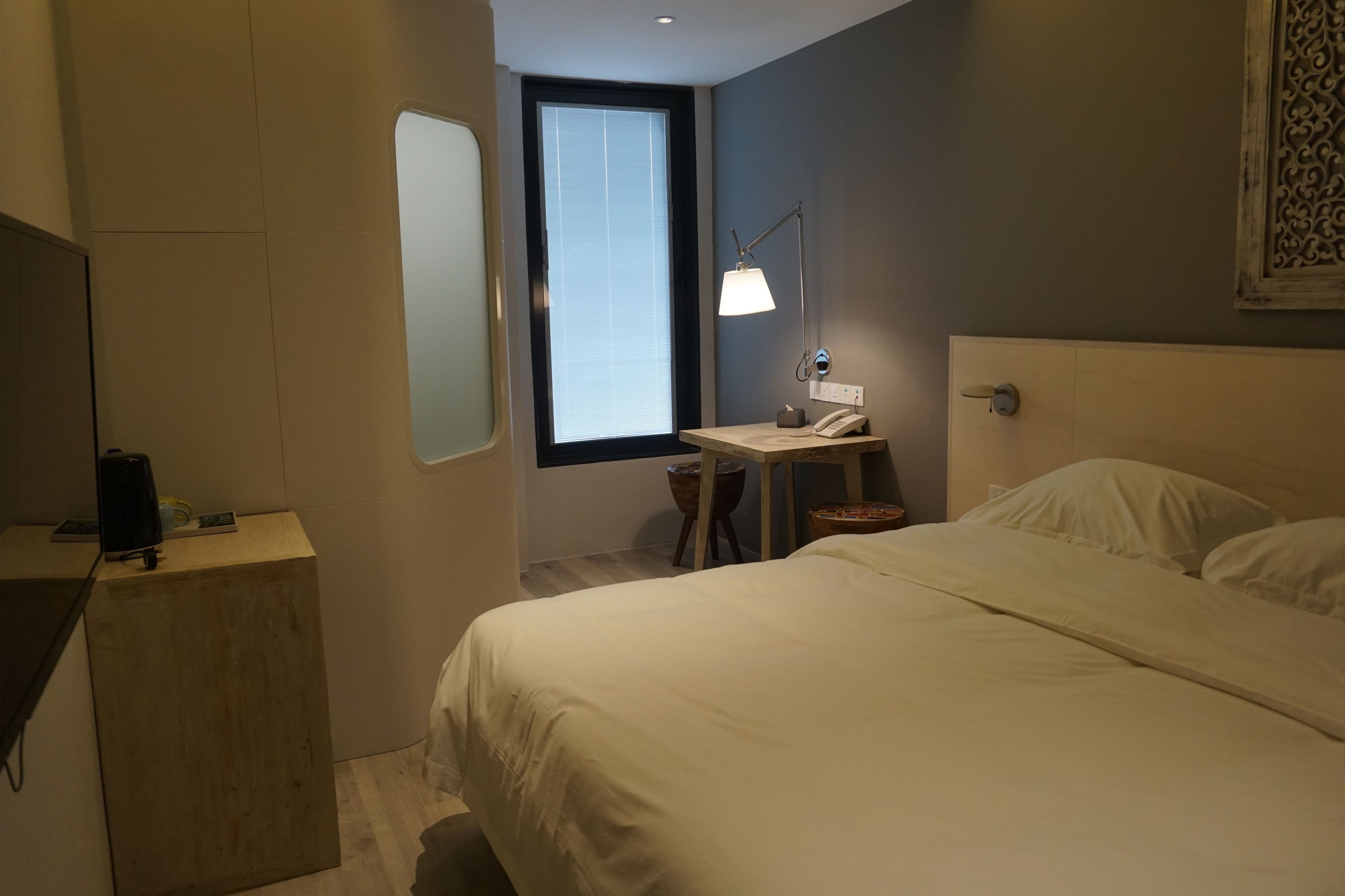 A hotel room with a bed, a desk with a lamp, a telephone, and a window with blinds.