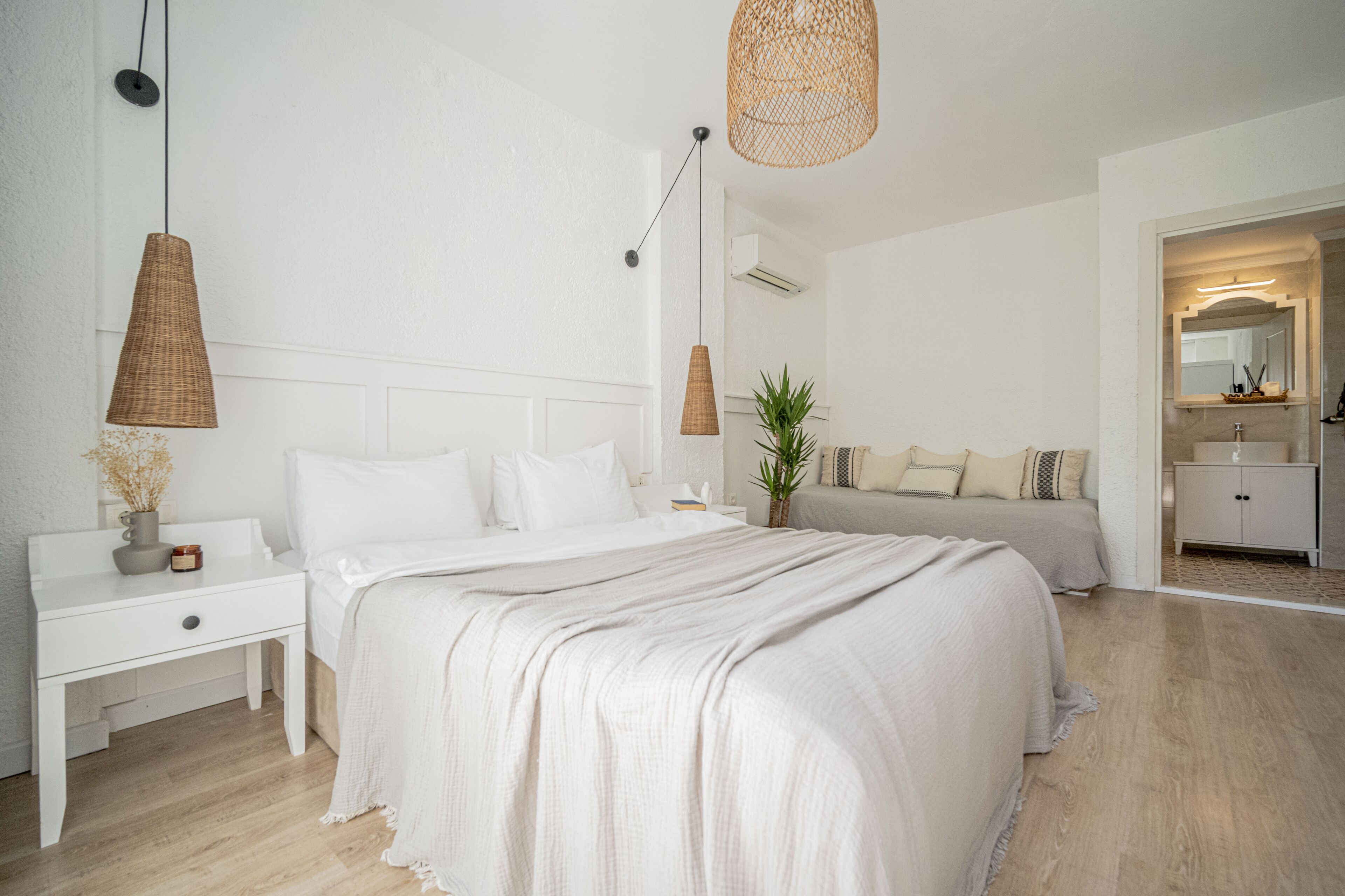 A bedroom with a large bed, bedside tables, a nightstand, a hanging light, a plant, and a bathroom visible through an open door.