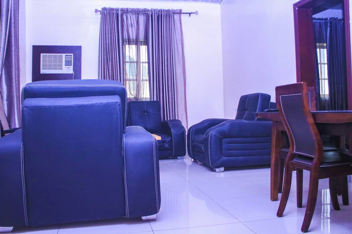 A room with a large blue sofa, a smaller blue armchair, a wooden dining table with chairs, and a window with curtains.