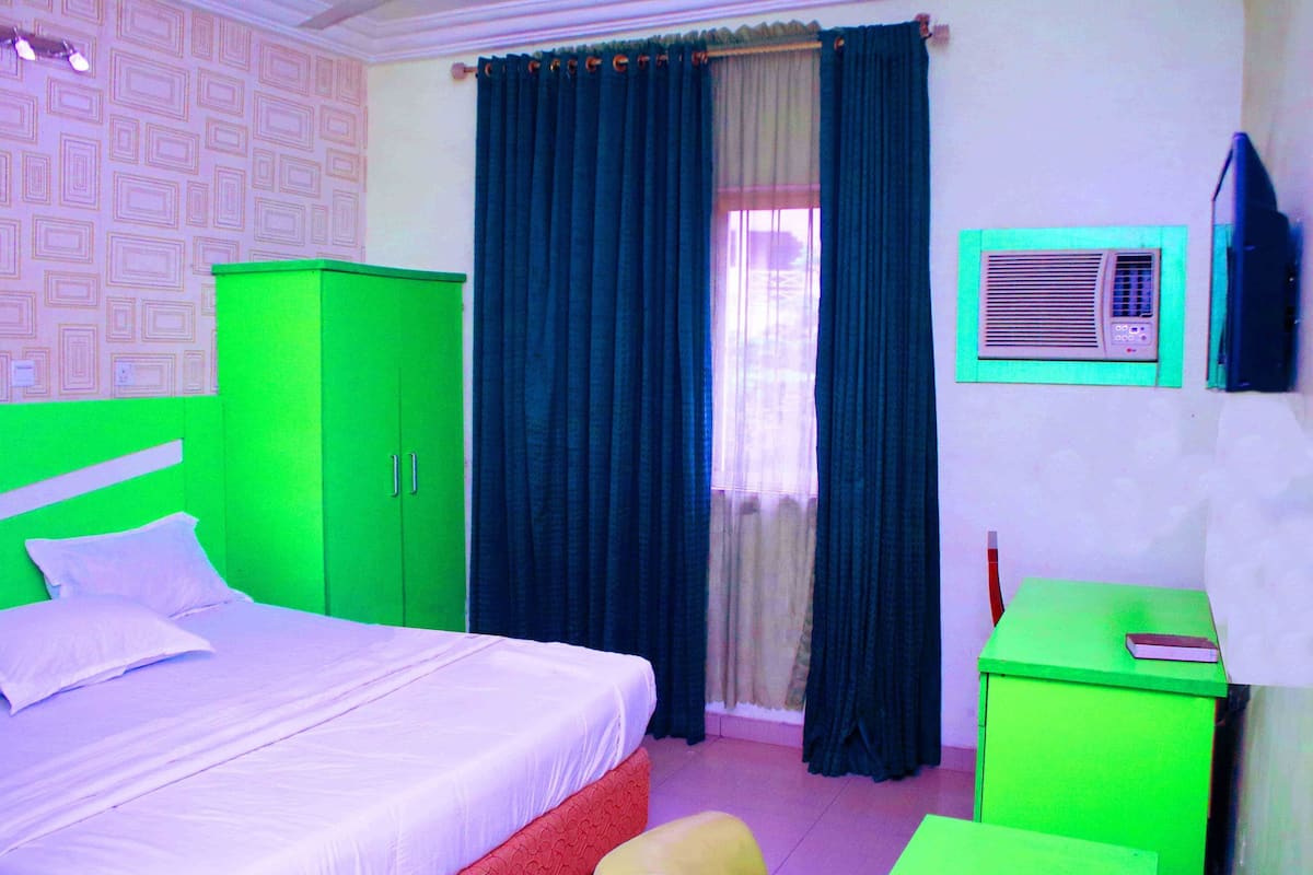 A room with a bed, a green wardrobe, a small table, a green cabinet, and a window with curtains.