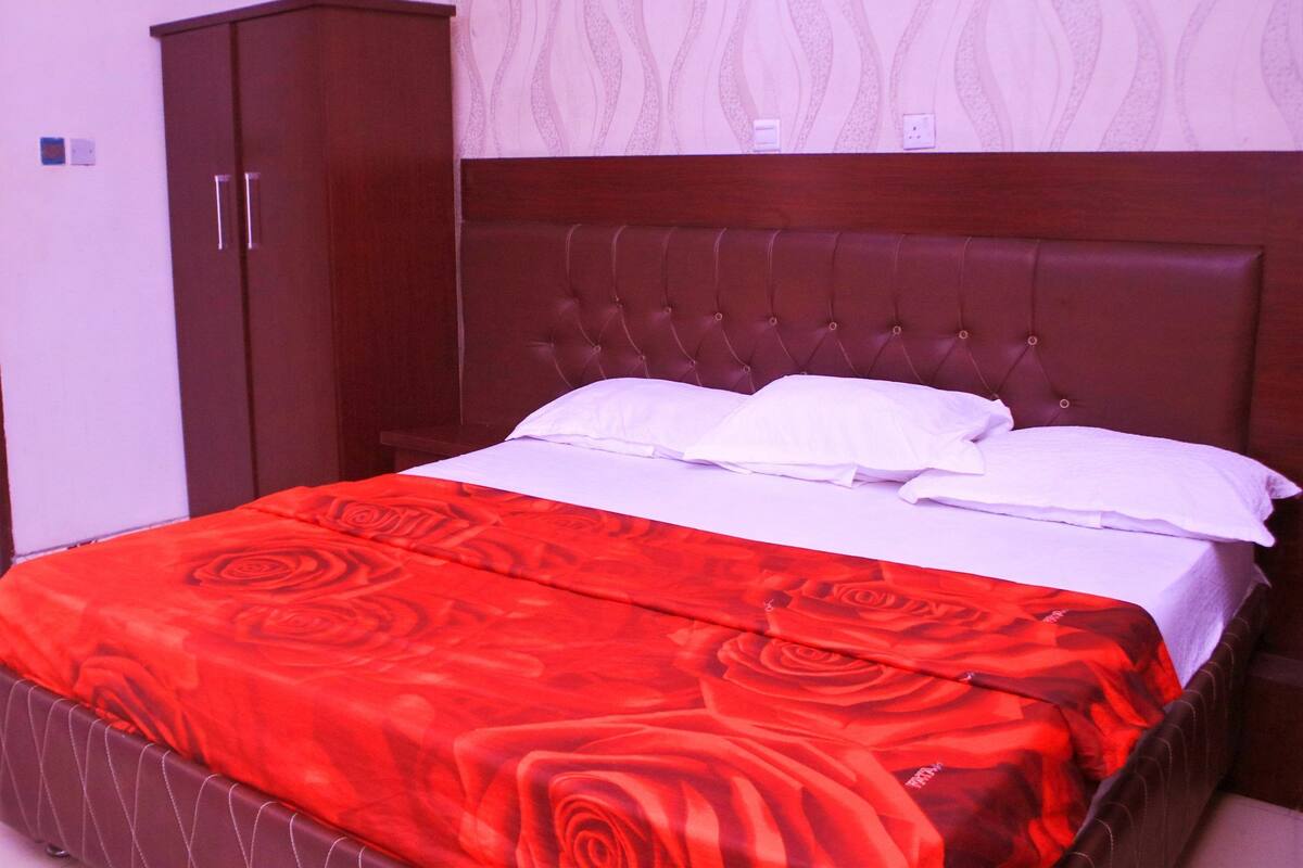 A neatly made bed with a red floral bedspread and white pillows, a wooden headboard, and a wardrobe to the left.