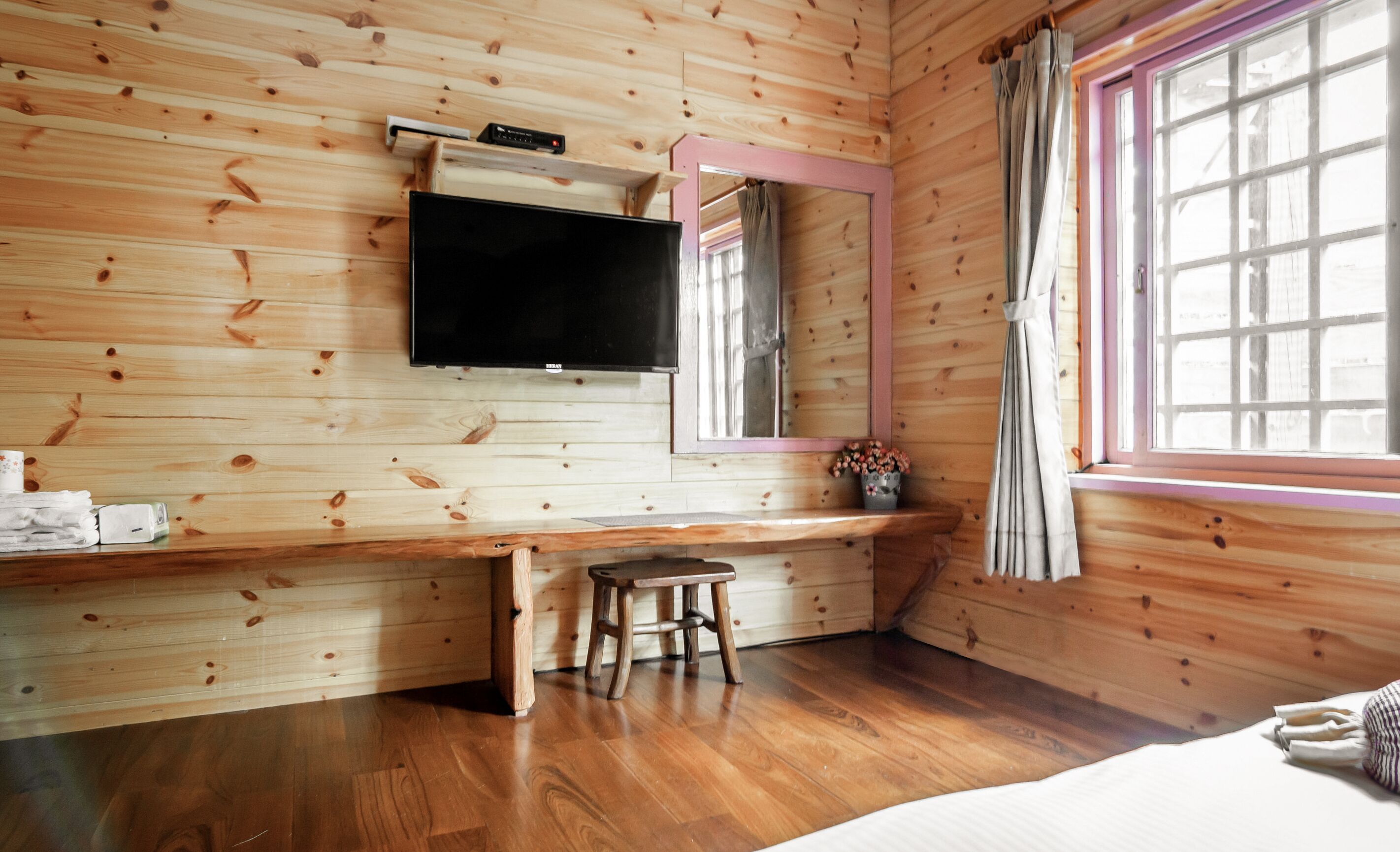 A wooden cabin room with a bed, a desk, a TV mounted on the wall, and a window with curtains.