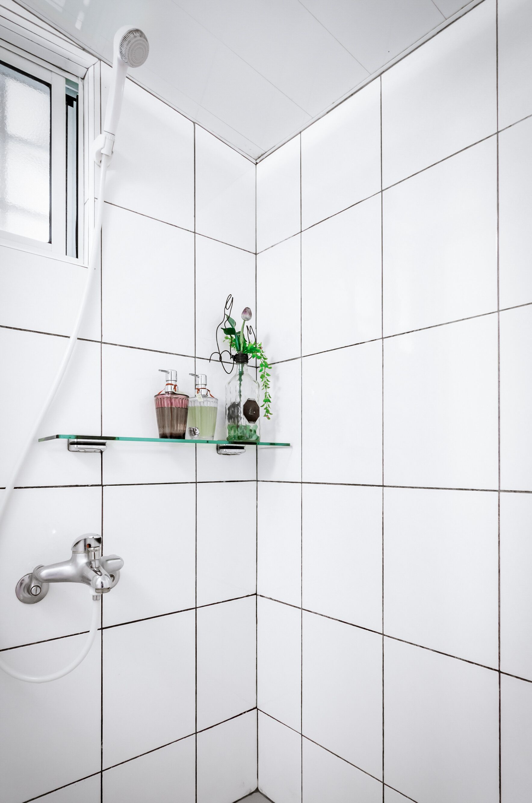 A bathroom with white tiled walls, a glass shelf with toiletries, a handheld showerhead, and a wall-mounted faucet.