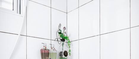 Un baño con paredes de azulejos blancos, un estante de vidrio con artículos de aseo, un cabezal de ducha manual y un grifo empotrado.