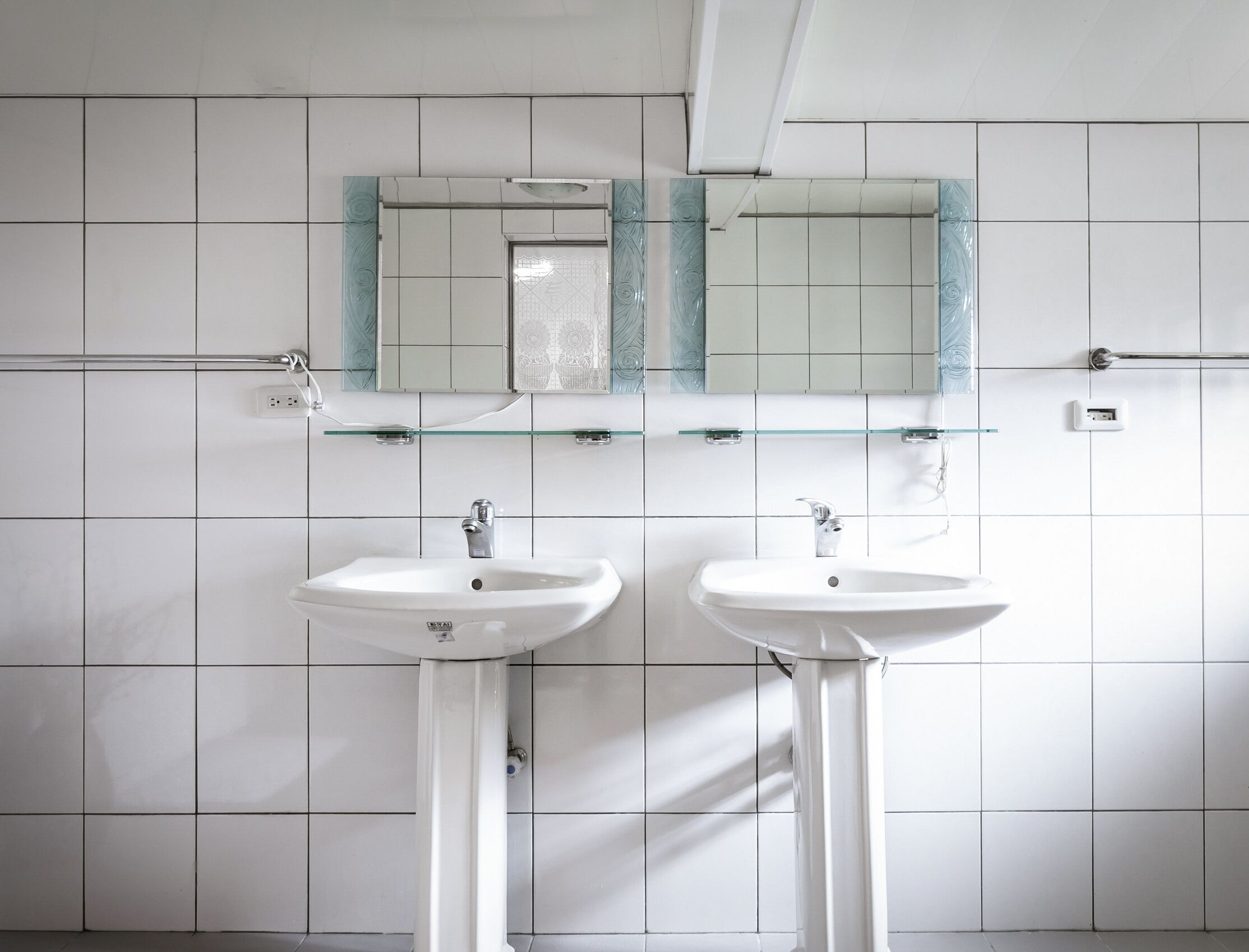 Un baño con dos lavamanos blancos, un espejo con marco de vidrio y paredes cubiertas de azulejos blancos.