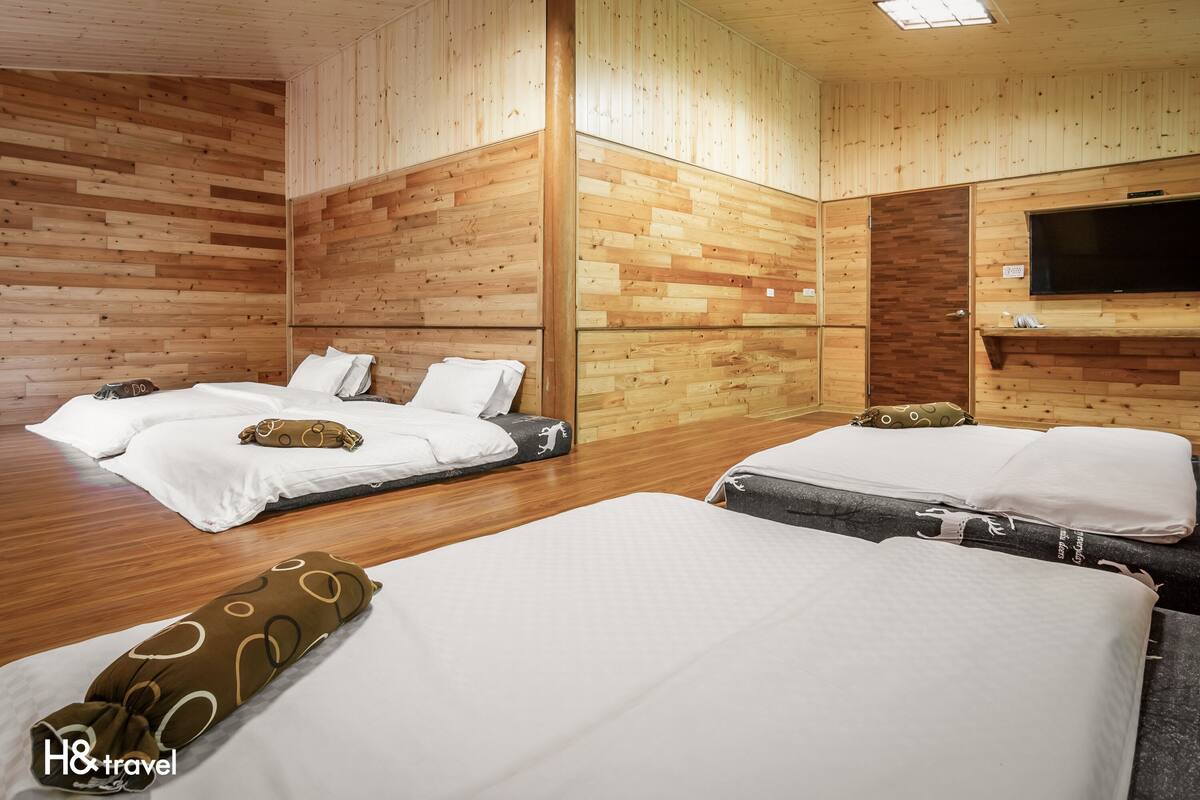 Habitación de hotel con tres camas, paredes de madera, un televisor de pantalla plana y suelo de madera.