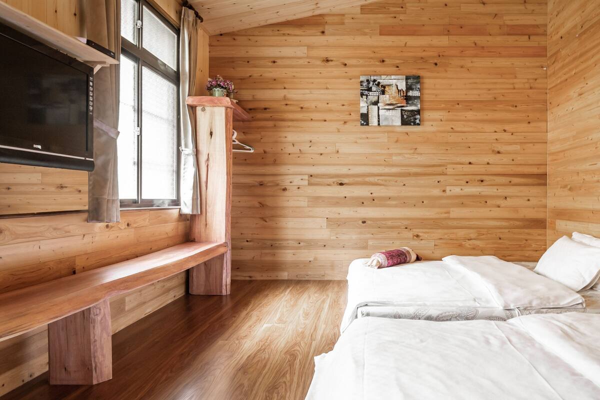 Una habitación con paneles de madera, una cama, un banco, un televisor y una ventana con cortinas.