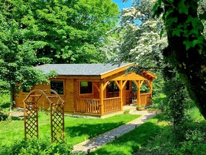 Une cabane en bois dotée d’un porche, entourée d’une végétation luxuriante et d’arbres.