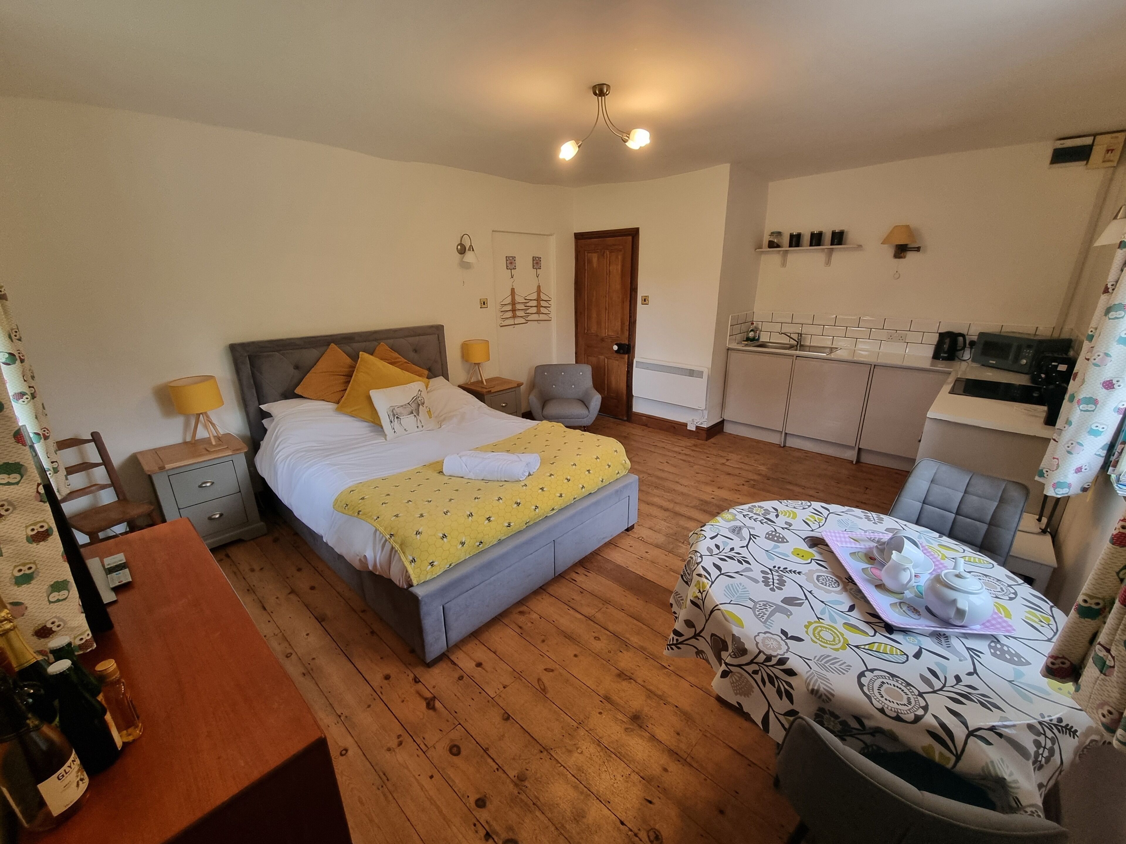 Ein gemütliches Schlafzimmer mit einem Bett, Nachttischen, einem kleinen Schreibtisch, einem Stuhl, einem Tisch mit blumigem Tischläufer und einer kleinen Küchenzeile im Hintergrund.