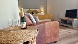 A cozy living room with a round wicker table, two wine glasses, and a bottle of wine. A brown leather couch and a television are also visible.