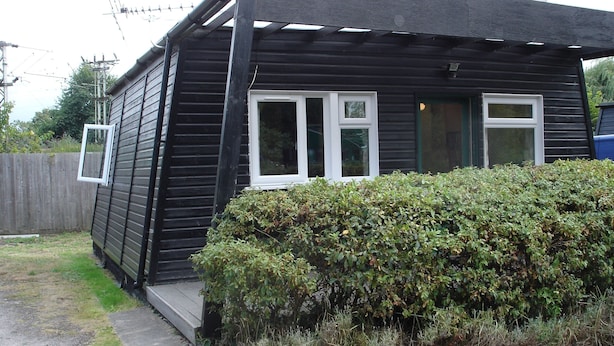 A small, single-story wooden house with a porch, a fenced yard, and a bush in front. - Broxbourne Riverside Chalets (Broxbourne)