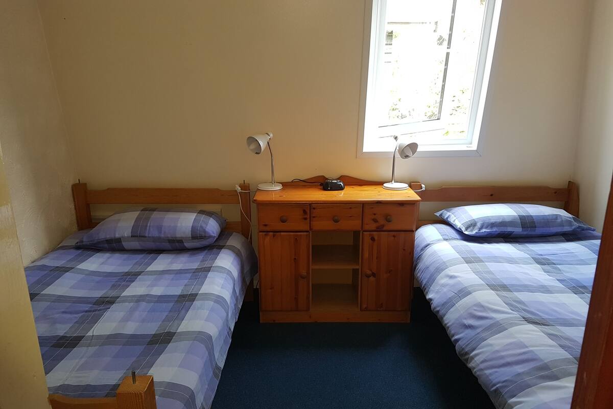 A small, single-room accommodation with two beds, a wooden desk, and a window providing natural light.