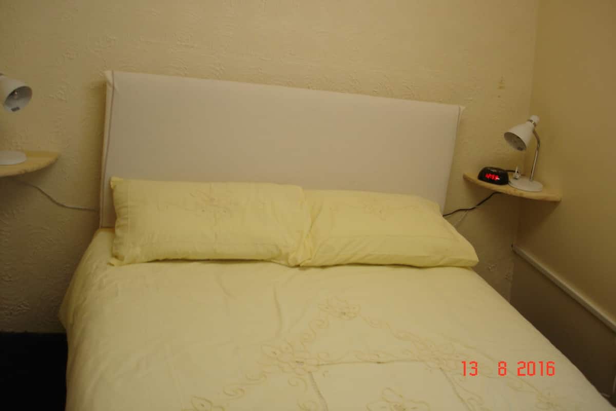 A small, single-bed room with a plain headboard, a bedside table with a lamp, and a wall clock.