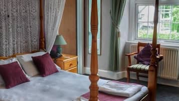 A four-poster bed with a canopy, a nightstand with a lamp, a chair, and a window with curtains.