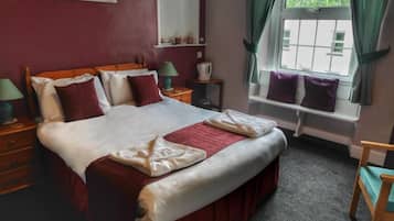A hotel room with a large bed, a window with curtains, a bedside table with a lamp, and a chair.