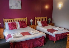 Habitación de hotel con dos camas, cada una con cabecero de madera, ropa de cama roja y sábanas blancas. Hay mesitas de noche con lámparas y una l ámpara de pared.