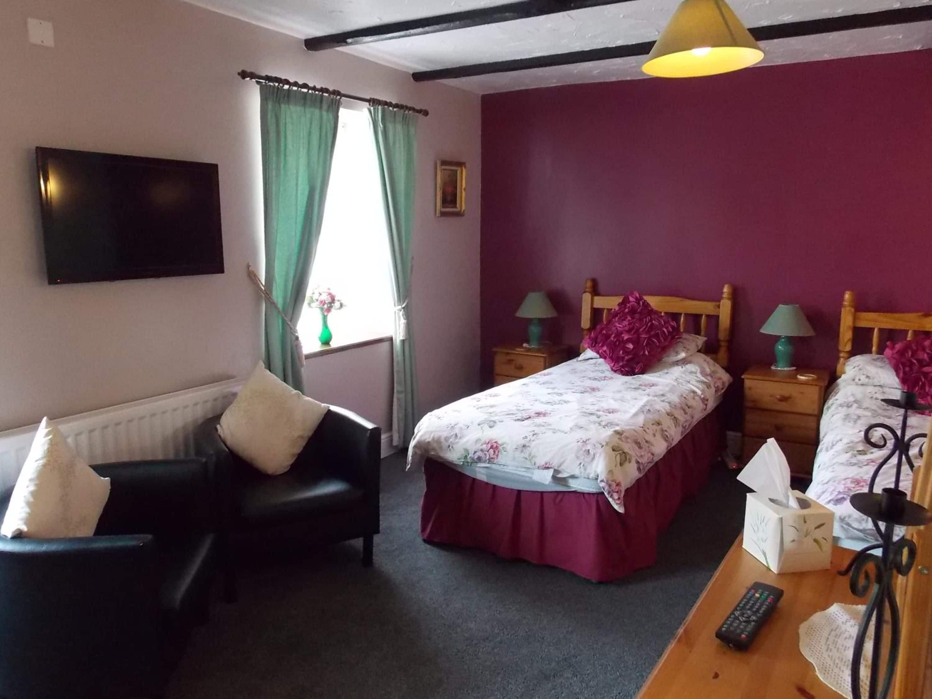 Un dormitorio con cama, una silla, mesita de noche, lámpara, televisor y ventana con cortinas.