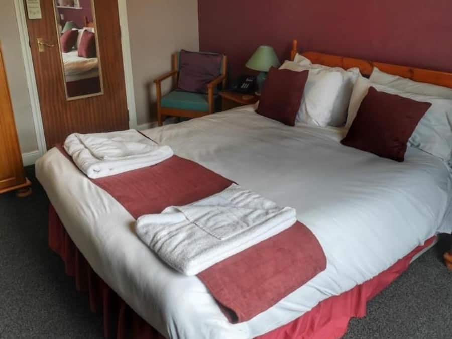 Quarto de hotel com uma cama grande, cabeceira de madeira, mesa de cabeceira com abajur, uma cadeira e um espelho.