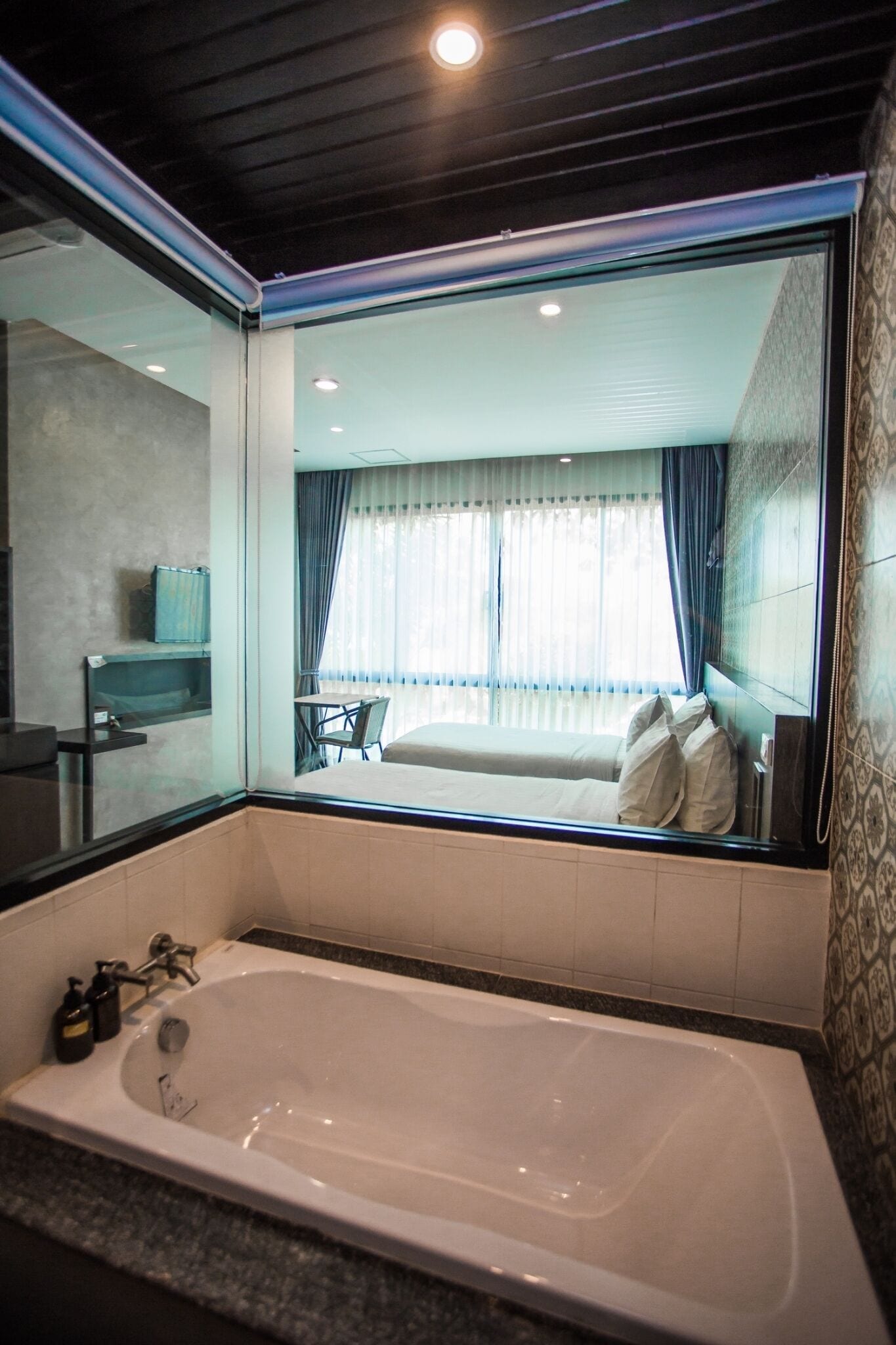 A modern bathroom with a large bathtub, a mirror, and a view of a bedroom with a bed and curtains.