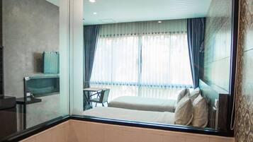 A modern bathroom with a large bathtub, a mirror, and a view of a bedroom with a bed and curtains.