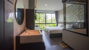 A modern hotel room with a large bed, a bathroom with a sink and mirror, and a view of greenery through large windows. - Anantaya Home Phare (Sung Men)