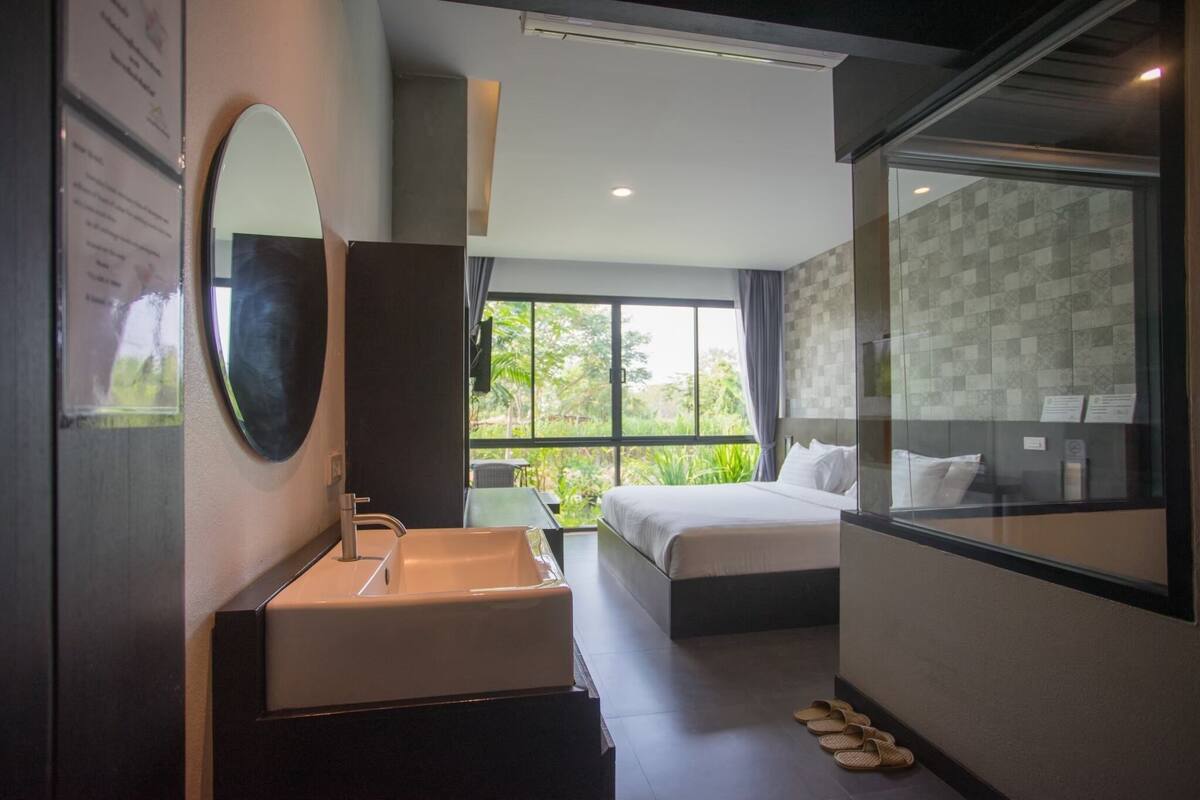 A modern hotel room with a large bed, a bathroom with a sink and mirror, and a view of greenery through large windows.