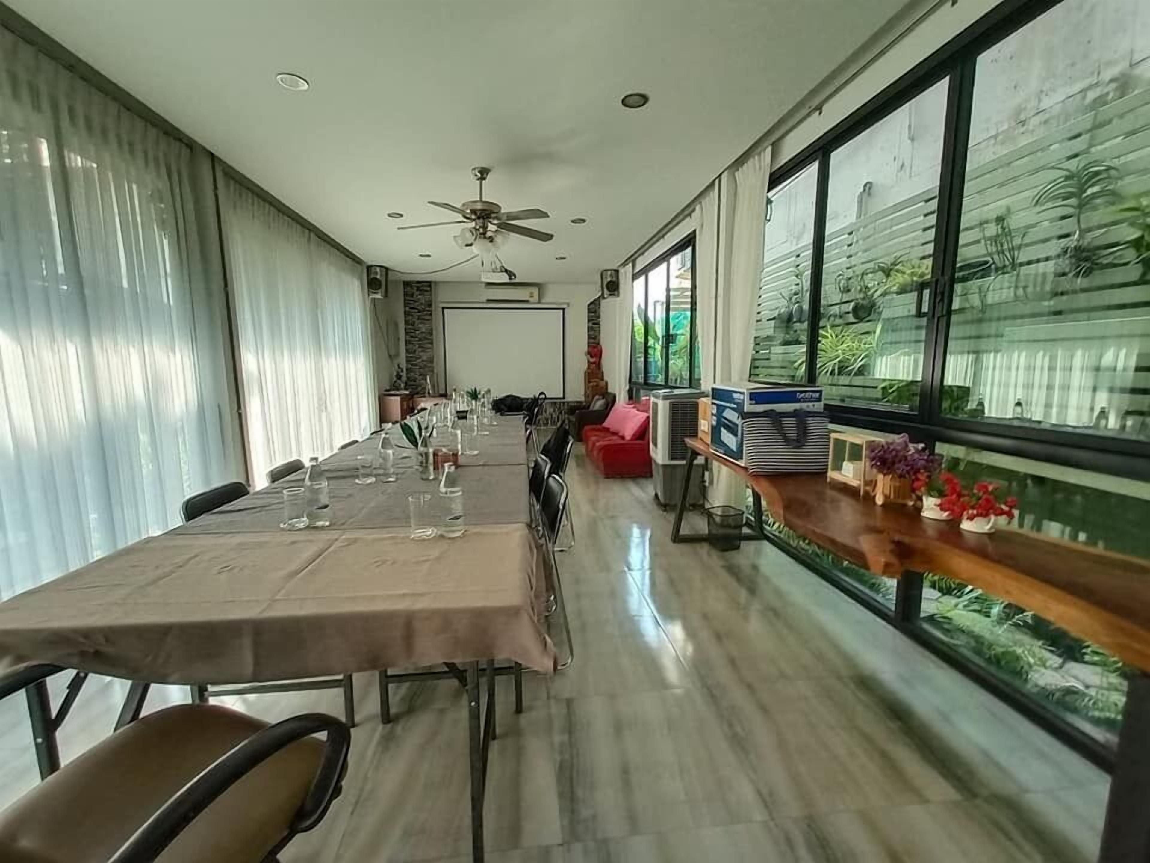Een lange eettafel gedekt voor een maaltijd, met stoelen aan weerszijden. Er is een plafondventilator, grote ramen met planten en een stenen muur op de achtergrond.