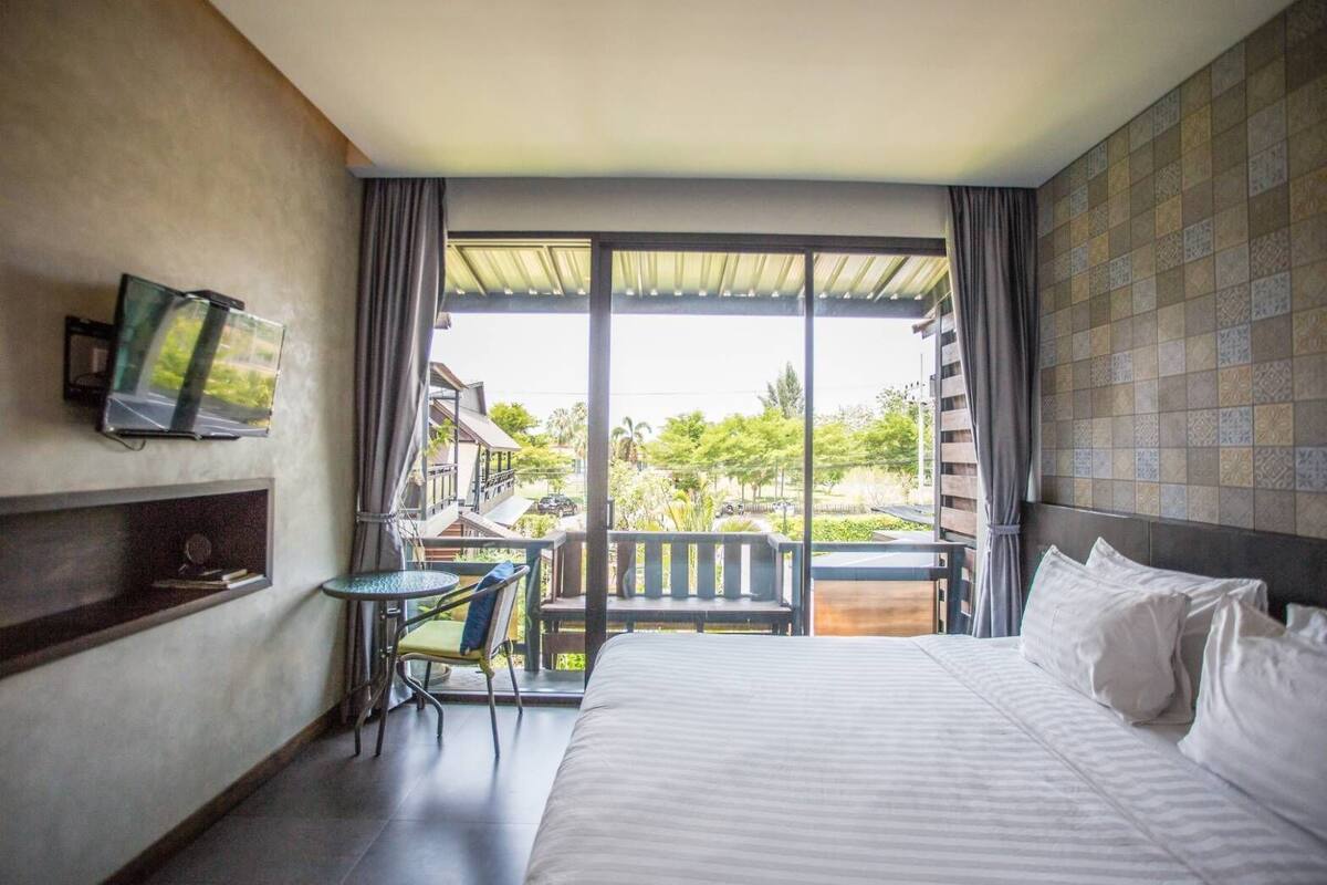 A hotel room with a bed, a small table with a chair, a TV mounted on the wall, and a balcony with a view of the outside.