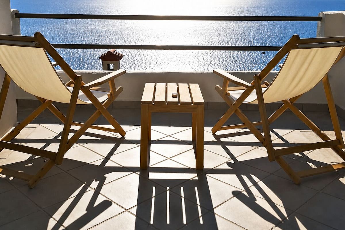 A balcony with two wooden chairs and a small table, overlooking a calm sea and distant mountains.