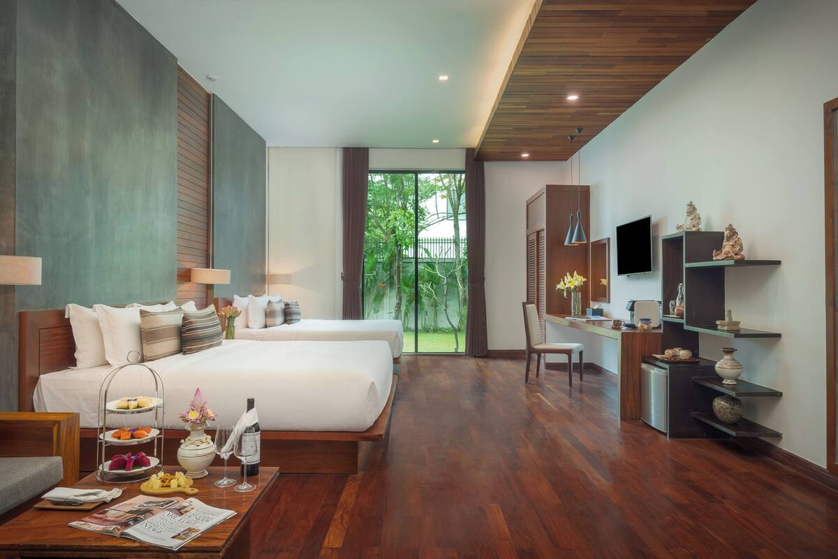 A modern hotel room with two beds, a dining area, a small kitchen, and a large window offering a view of greenery.