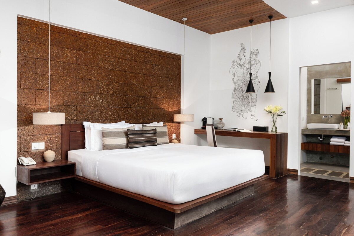 A modern hotel room with a large bed, wooden headboard, and a stone accent wall.