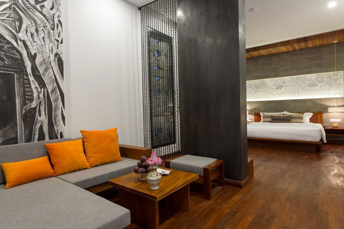 Una habitación de hotel moderna con un sofá gris, cojines anaranjados, una mesa de centro de madera con un jarrón y una cama con cabecera blanca.