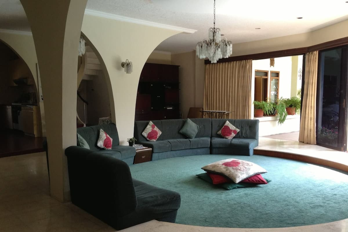 A spacious living room with a large sectional sofa, a small coffee table, and a chandelier. There is a sliding glass door leading to a balcony with potted plants.
