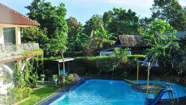 A swimming pool with a slide, surrounded by lush greenery and a thatched-roof structure. - The Garden Family Guest House (Ciawi)